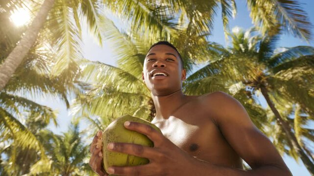 A happy young man with his arms around two coconuts standing in front of palm trees on a sunny day.