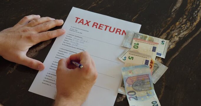 Person completing a tax return document with a pen while cash lies beside it, showing finance, paperwork and tax preparation.