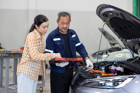 Auto mechanic guiding a customer while inspecting a car engine in a repair shop, using a digital tablet for diagnostics under the open hood.