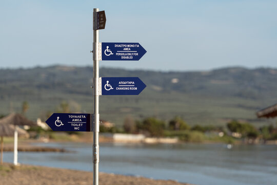 Blue wheelchair accessible signs in Greece pointing to toilet WC, changing room, and pergola only for disabled. Inclusive resort infrastructure directional arrows for handicap people on the beach.