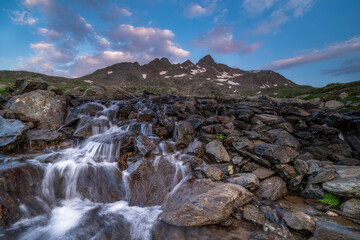 Fototapeta premium Long exposure of a cascading mountain stream with jagged peaks under a soft twilight sky, perfect for travel and environmental themes.