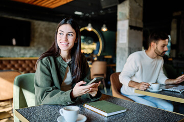 Man and woman smiling, holding coffee while working on a laptop and using a mobile phone  in a cafe
