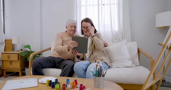 Slow motion senior couple browse tablet references beside paint supplies and easel, smiling and planning artwork before painting at home, expressing companionship, mindfulness, and healthy aging.