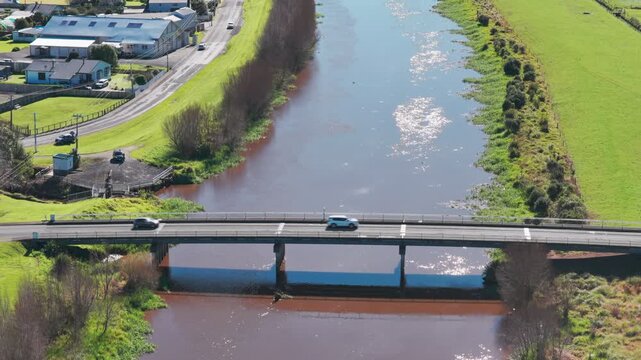 A car drives over a bridge spanning a wide, muddy river in a rural area of Ngatea, New Zealand. The sun glints off the water's surface.