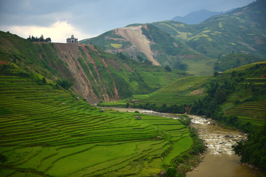 Terraced Rice Fields and River: Verdant, meticulously sculpted rice terraces cascade down the hillsides, bordering a winding river that flows through a lush valley under a dramatic sky.