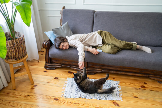 Happy relaxed woman lying on couch in living room playing with cat, peaceful lazy hours at home, smiling female procrastinating time on sofa enjoying interactions with pet, laziness and comfort.