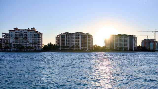 The sun sets behind rows of waterfront high rise buildings reflecting on the calm bay