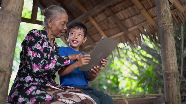 Elderly woman leans beside smiling young boy as both focus on handheld tablet inside rustic open-air bamboo shelter, with woven thatch roof, rough wooden posts, soft daylight, and calm rural