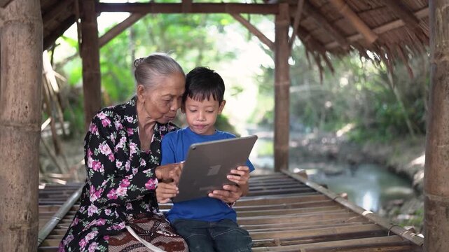 Elderly woman and young boy sit close together on bamboo platform inside open rustic shelter, sharing tablet with focused smiles, soft daylight, green background, and calm village stream beside
