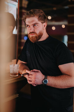 A bearded man leans at a bar holding a cigar and a glass of whiskey. He appears thoughtful and relaxed in a dim, modern lounge setting.