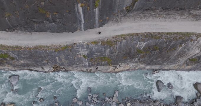Annapurna Circuit, Manang Nepal, top-down aerial tracking shot of a hiker walking on a narrow dirt route carved into a steep rock cliff face directly above the white water rapids of Marsyangdi River