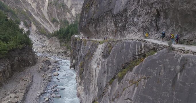 Annapurna Circuit, Manang Nepal, aerial glide shot of hikers walking on a narrow dirt ledge cut into a vertical rock cliff directly above the winding white water rapids of the Marsyangdi River gorge.
