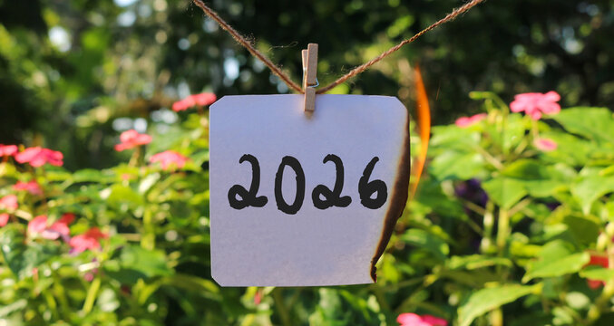 Burning paper with the text 2026 hanging on a rope in a colorful flower garden. End of 2026, say goodbye to 2026. New Year's concept 2027
