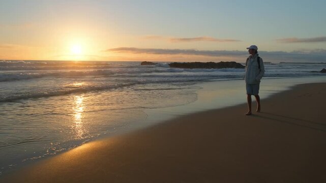 Happy young man walking on a sandy ocean beach on a sunny summer day. Young Male guy walking along the sandy coast on a sunny evening. A guy with a backpack walks on a beach