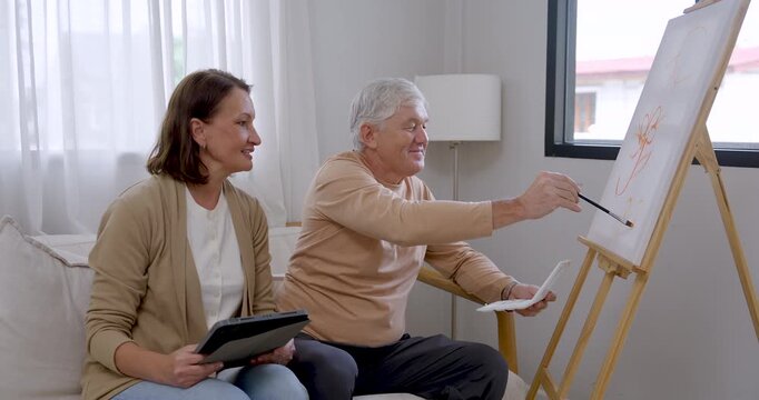 Senior man paints orange flower lines on canvas while senior woman smiles beside him with tablet, creating home art therapy scene for emotional wellness, relaxation, companionship, active retirement.
