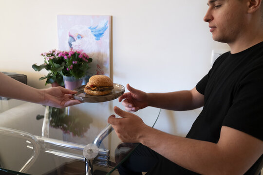 Man receiving a delicious burger on a plate from a woman in a cozy home setting with a colorful plant in the background.