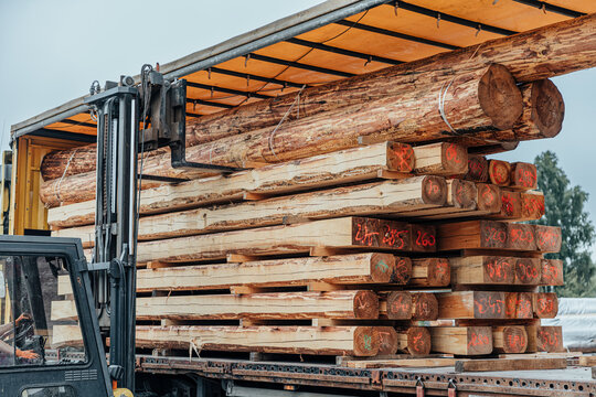 Forklift operator loading stacked rough-sawn timber beams and logs onto a cargo truck for export at a lumber yard. Marked wood ends and copy space are visible