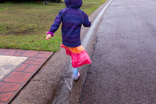 child in raincoat and boots splashing through roadside gutter full of water on rainy day