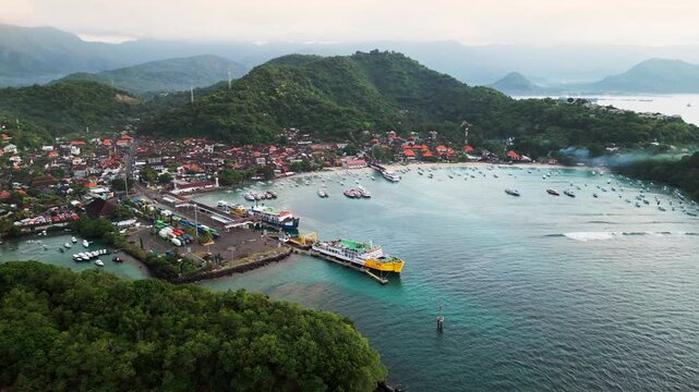 Passenger ferries docked at a busy ferry terminal beside a coastal town and tropical hills in Bali Indonesia, aerial drone footage.