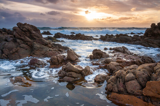 Low angled view of waves washing over rocks along a rugged coastline under a colourful sunset sky