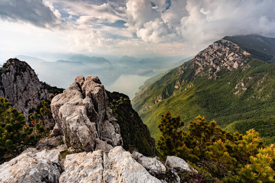 Rugged rocky cliffs and pine trees overlook the deep valley and Lake Garda, representing nature exploration at Ventrar trail, Monte Baldo Natural Park, Brentonico,Trentino,Veneto Italy.