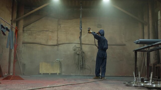 Industrial worker spraying paint onto suspended metal beams inside spacious workshop with protective coveralls, respirator, hoses, overhead lights, and visible mist