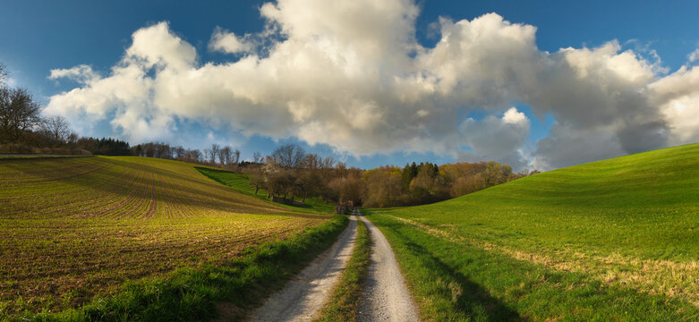 Rural dirt road in idyllic landscape with meadows, fields, hills and amazing clouds
