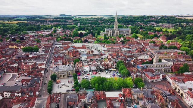 Wide drone view over Salisbury city centre in Wiltshire, showing the historic Market Square, Guildhall, city rooftops, streets, trees and Salisbury Cathedral spire in the distance.