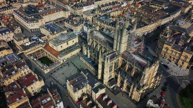 Close aerial drone footage over Bath, England, showing Bath Abbey's Gothic towers and buttresses alongside the Roman Baths courtyard, church square below, and surrounding honey-stone rooftops
