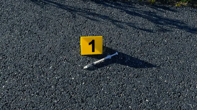 Yellow evidence marker number one placed next to a used syringe on the asphalt, marking a piece of evidence at an outdoor crime scene investigation related to drug abuse or overdose