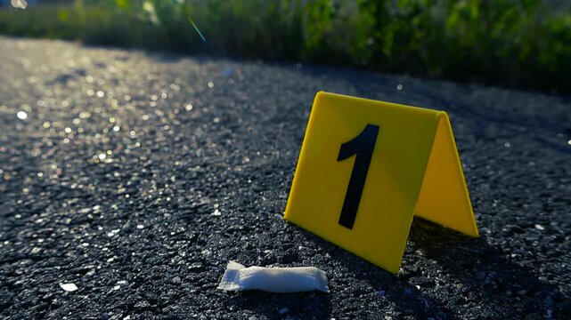Dramatic sunlight shift on a crime scene with a yellow evidence marker number one next to a white nicotine pouch, or snus, lying on the asphalt pavement with green grass in the background
