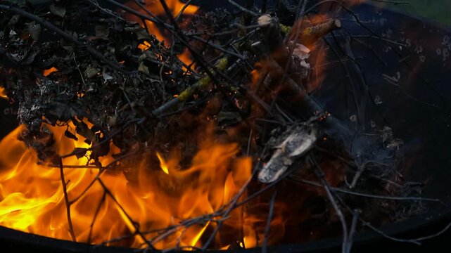Close up of garden waste, including dry leaves and wooden branches, burning intensely inside a dark metal incinerator bin, with bright orange flames consuming the brushwood
