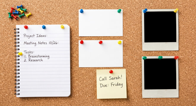 Brown cork board displays a spiral notebook with handwritten meeting notes alongside several blank white cards and empty polaroid picture frames held in place by colorful plastic push pins everywhere