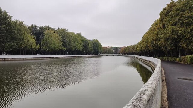 The river flows through the city among lawns where chestnuts, spruce and linden trees grow, reflected in the water. The riverbanks are concrete. There is a parapet and asphalted path nearby. Cloudy