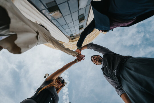 A diverse business team stands outdoors and joins hands above their heads in a team gesture.