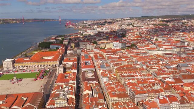A drone glides above a historic plaza and waterfront boulevard toward a distant suspension bridge