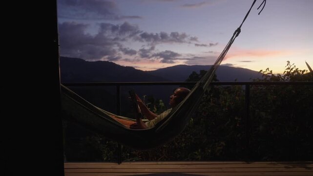 A man reading a book while lying in a hammock on a terrace surrounded by nature, with a spectacular sunset