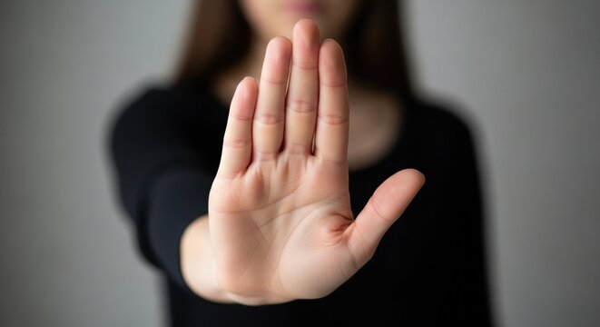 Woman holding hand up in a stop gesture. Female palm showing halt or rejection sign on blurred grey background
