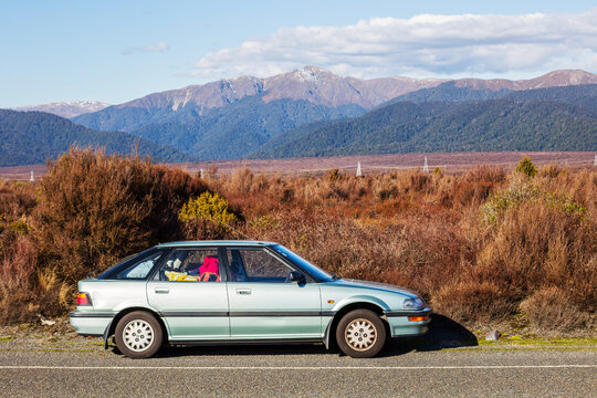 An older blue‑green sedan rests on the roadside of Desert Road, New Zealand, surrounded by dry alpine plains and distant snow‑tipped mountains under a clear sky