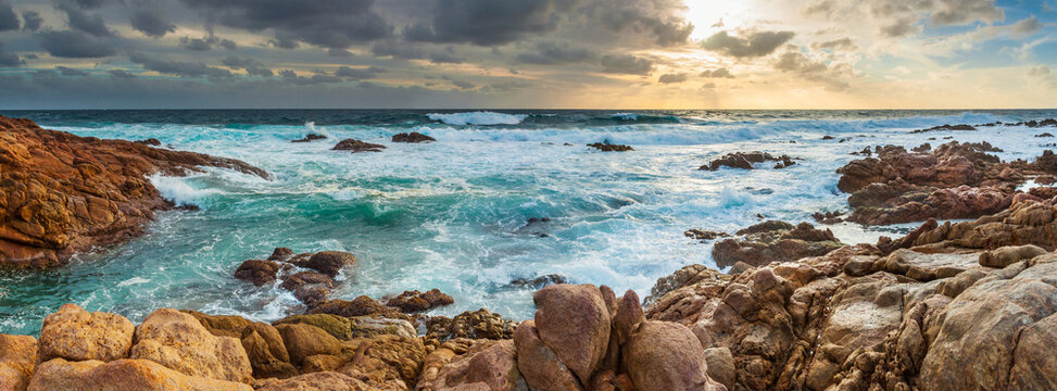 Panorama view of waves washing over rocks along a rugged coastline under a colourful sunset sky