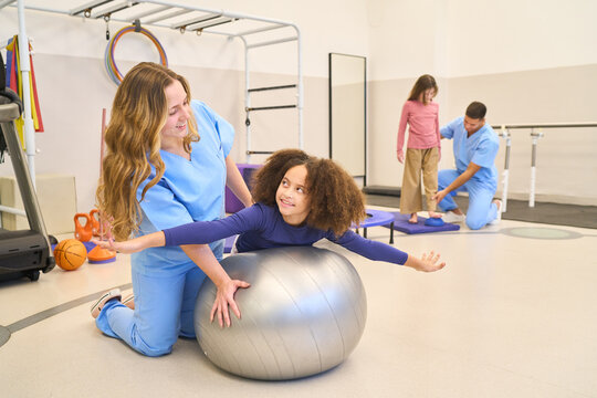 Pediatric physiotherapy session, therapist assisting child with balance exercises on fitness ball, rehabilitation exercises for motor skills training in clinic setting