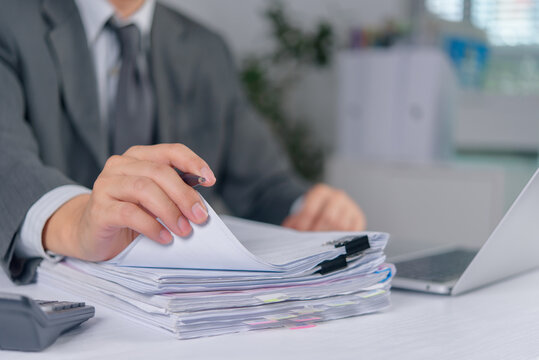 Businessman in a dark suit holds a pen while reviewing a massive stack of paperwork at an office desk. The professional auditor manages heavy administrative workload with a laptop and calculator.