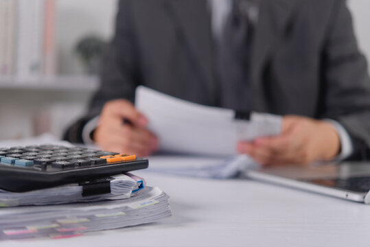 Black calculator rests on a thick stack of paperwork on a desk. In the blurred background, an accountant in a suit reviews financial documents during a heavy corporate auditing and budget planning.