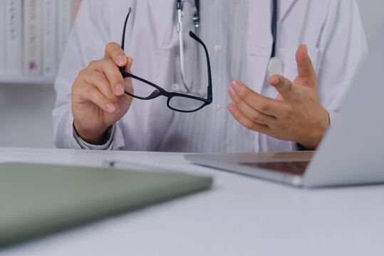 Healthcare professional in white coat holds black eyeglasses while gesturing during a consultation. The physician gives medical advice and explains diagnosis at a modern clinic desk with laptop.