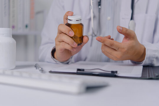 Doctor in white lab coat holds a brown medicine bottle and gestures with hand. Medical professional explains prescription dosage and pharmaceutical treatment during a clinical desk consultation.