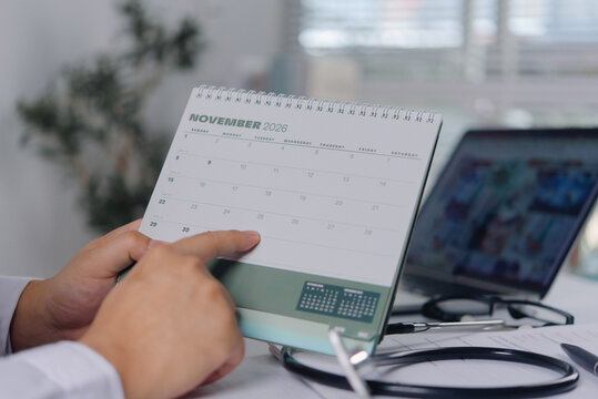 Doctor in a white coat points at a November 2026 desk calendar to schedule a medical appointment. The physician plans treatment dates and health checkups at a clinic workspace with a computer.