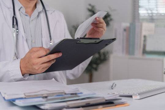 Medical professional in white lab coat and stethoscope reviews patient records on a clipboard. The focused doctor reads clinical documents at a desk in a bright, modern healthcare clinic office.