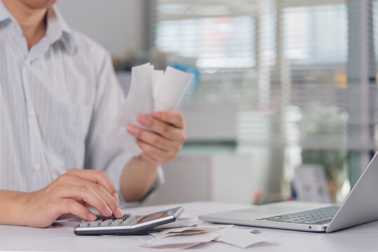 Man in a shirt uses a black calculator while holding paper receipts at a desk. The person manages personal finances, calculates household budget, and tracks monthly expenses next to a silver laptop.