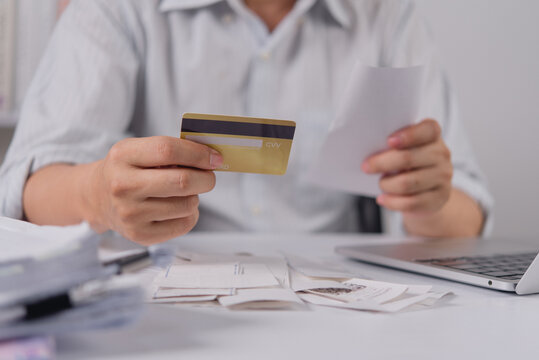 Hands of a man holding a gold credit card and a printed receipt at a desk. The person reviews monthly expenses and financial bills surrounded by a messy pile of paper receipts and a modern laptop.