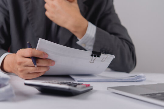 Stressed businessman in a suit holds a pen and touches his neck while reviewing a challenging document. The corporate professional analyzes difficult financial reports at an office desk workspace.